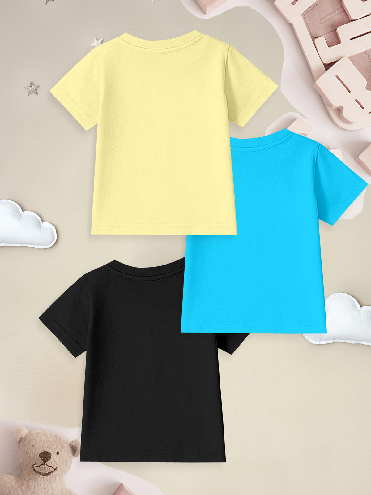 Three plain kids' t-shirts in yellow, blue, and black on a soft beige background with toys.