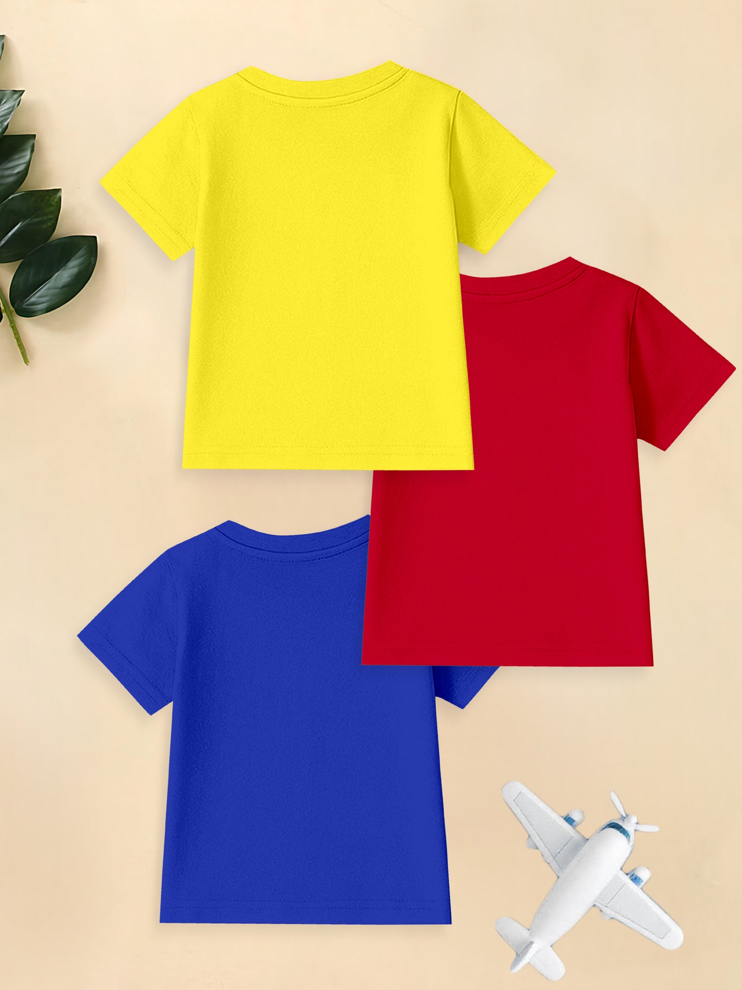 Three plain t-shirts in yellow, red, and blue with toy airplane and green leaves on beige background