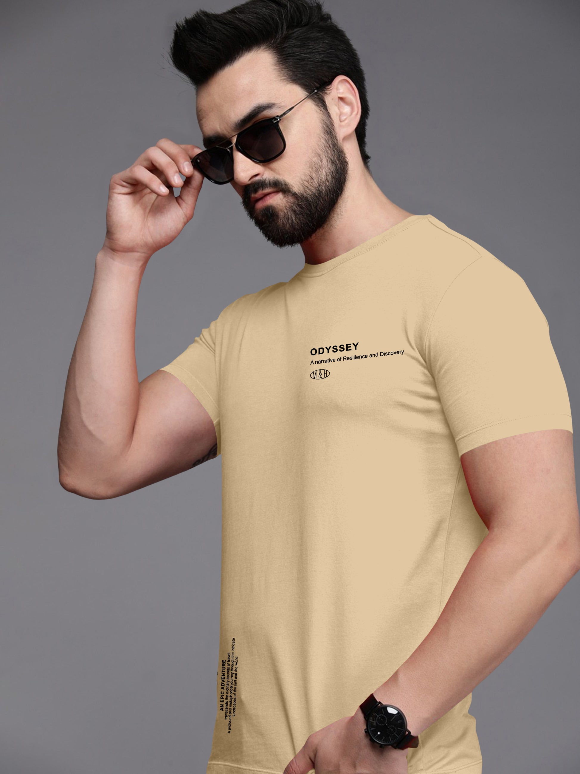 Man wearing beige graphic t-shirt with 'Odyssey' text, sunglasses, and wristwatch