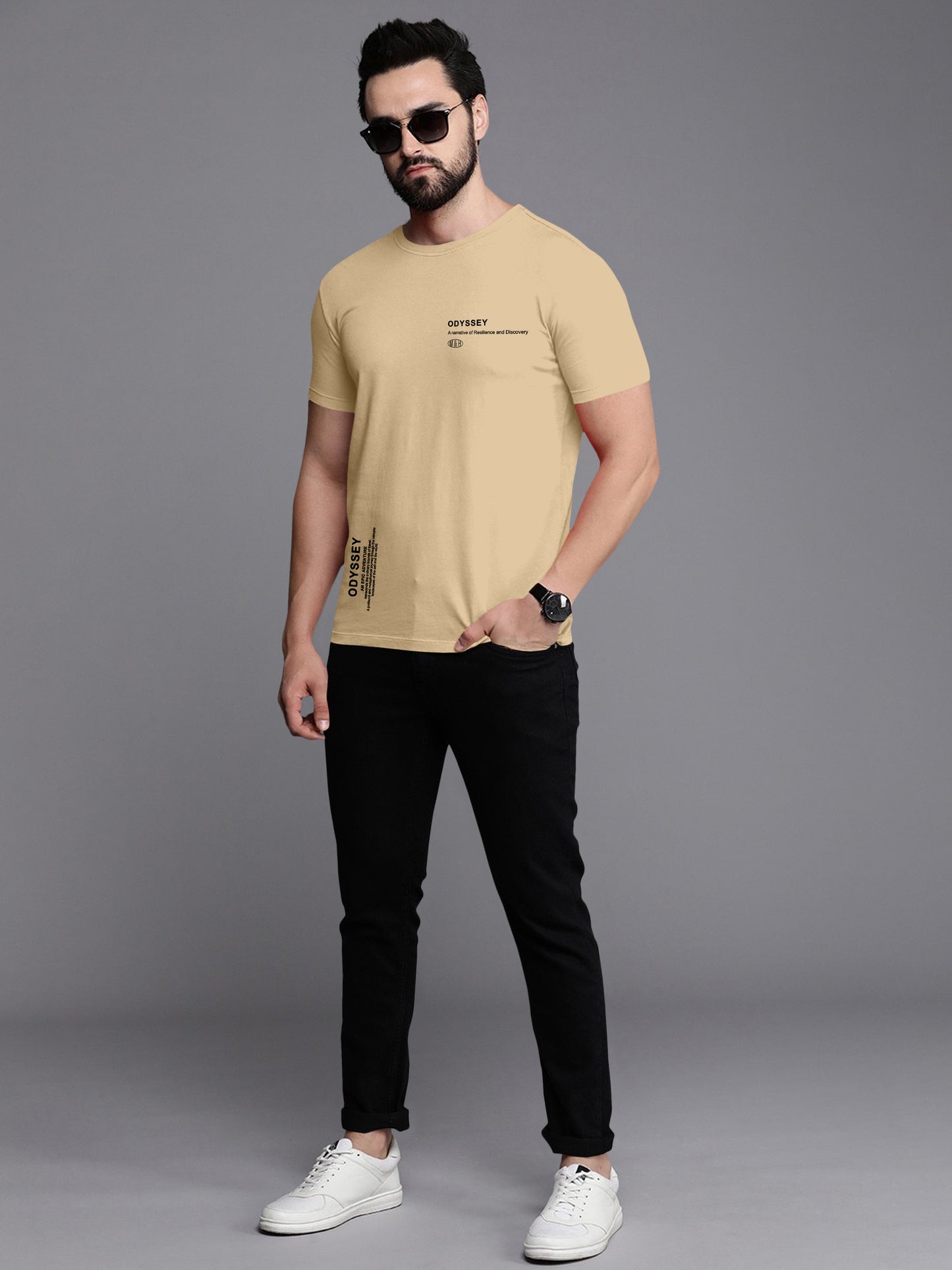 Man wearing beige graphic t-shirt with black text, black jeans, white sneakers, and sunglasses