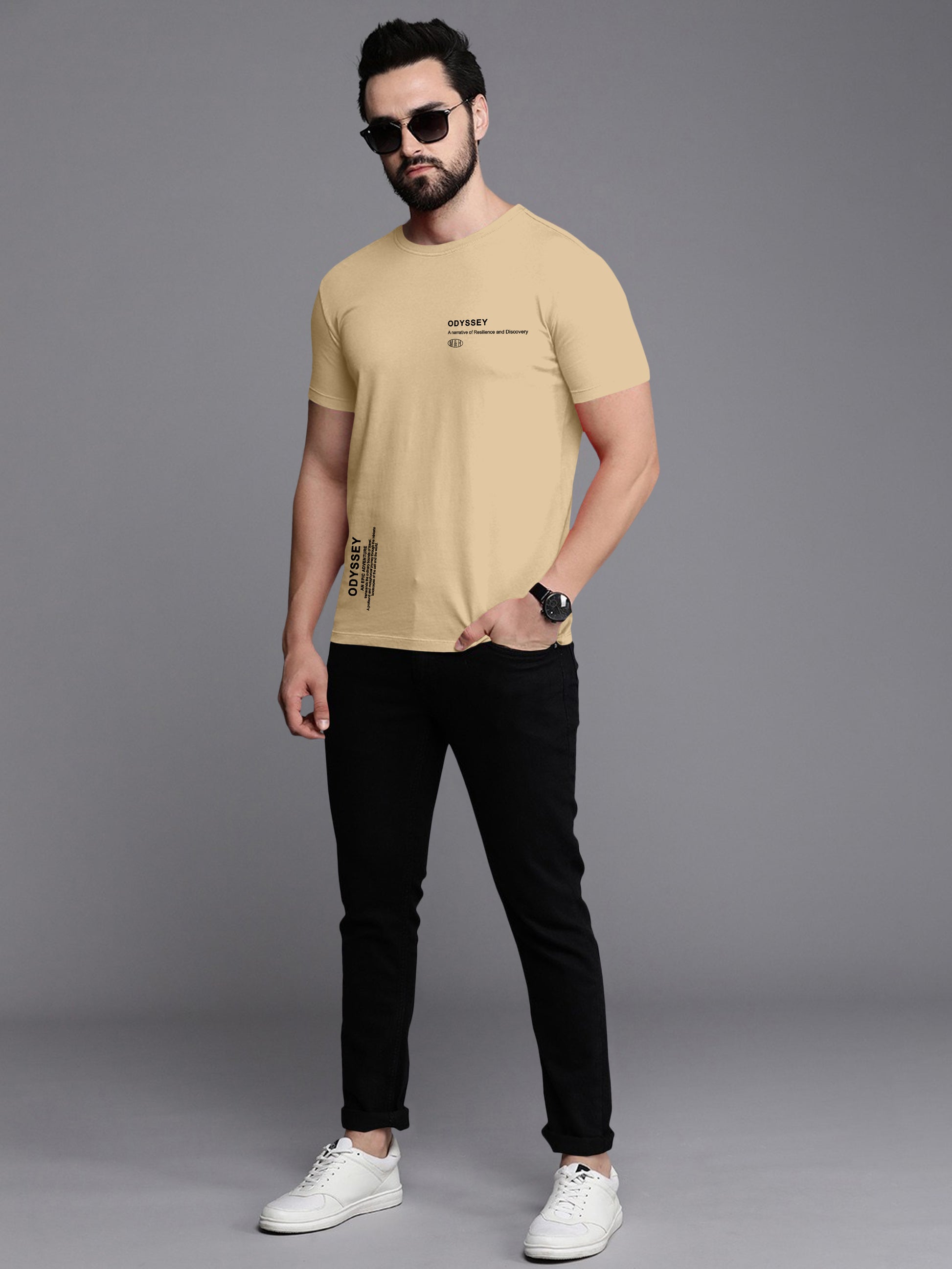 Man wearing beige graphic t-shirt with black text, black jeans, white sneakers, and sunglasses