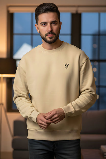 Man wearing a beige crewneck sweatshirt with small black chest logo, standing indoors
