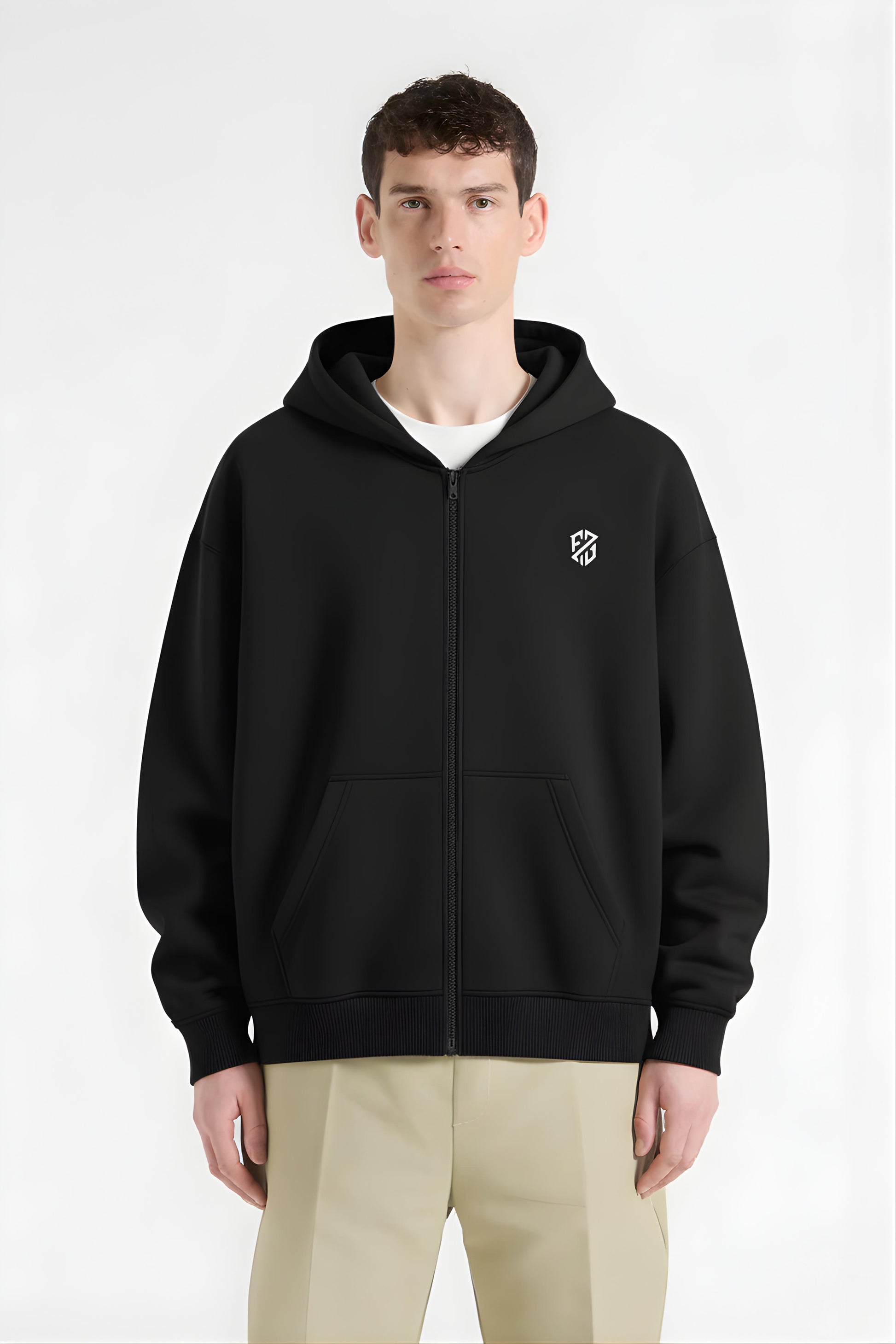 Man wearing a black zip-up hoodie with white chest logo and khaki pants on plain background.