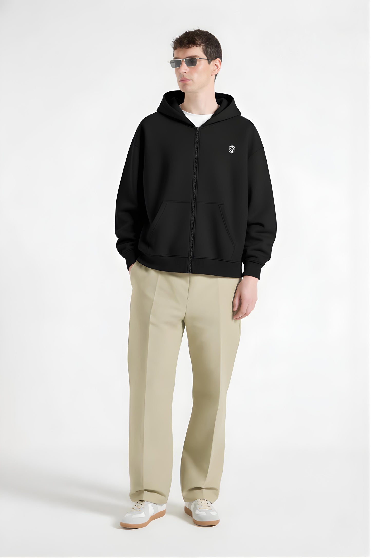 Man wearing black zip-up hoodie with logo, beige trousers, white sneakers, and sunglasses