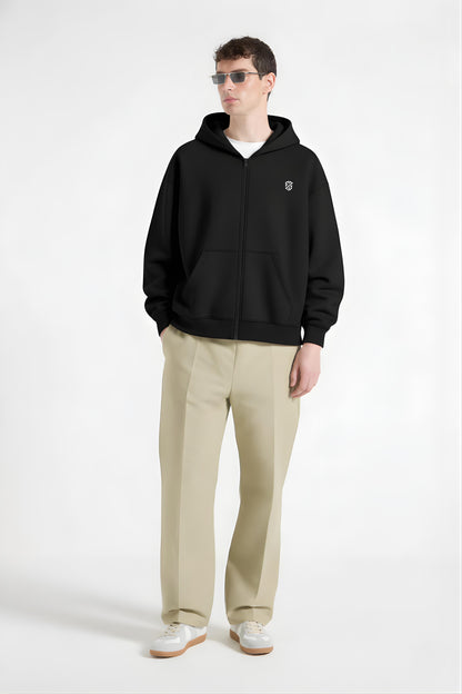 Man wearing black zip-up hoodie with logo, beige trousers, white sneakers, and sunglasses