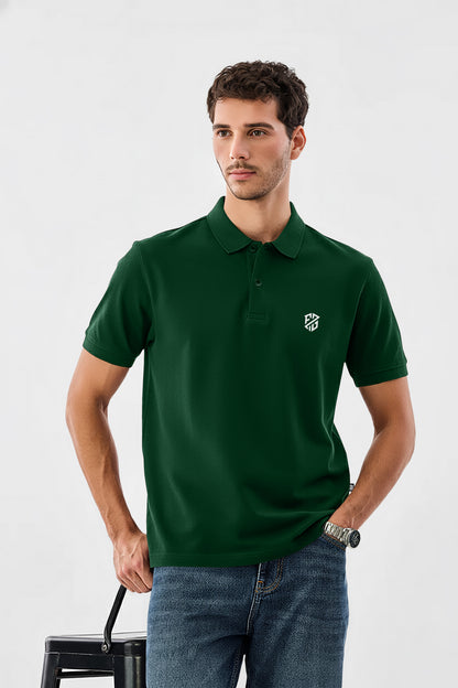 Man wearing dark green polo shirt with logo, blue jeans, and watch, standing indoors