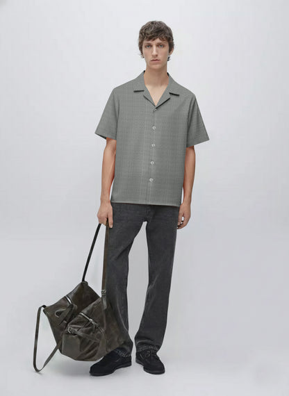 Man in short sleeve patterned shirt, black jeans, holding large dark shoulder bag