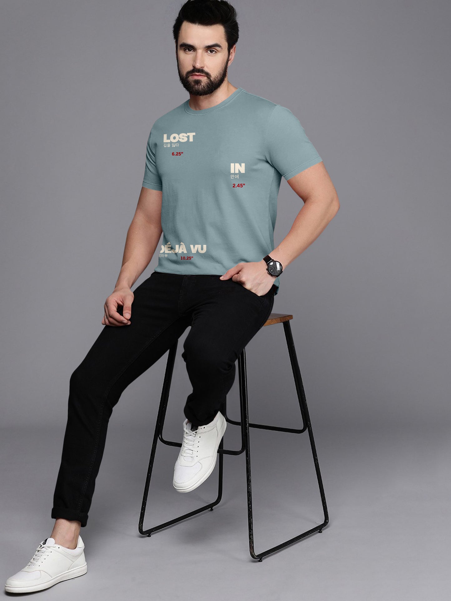 Man in blue graphic t-shirt with 'Lost in Déjà Vu' print, black jeans, white sneakers, stool