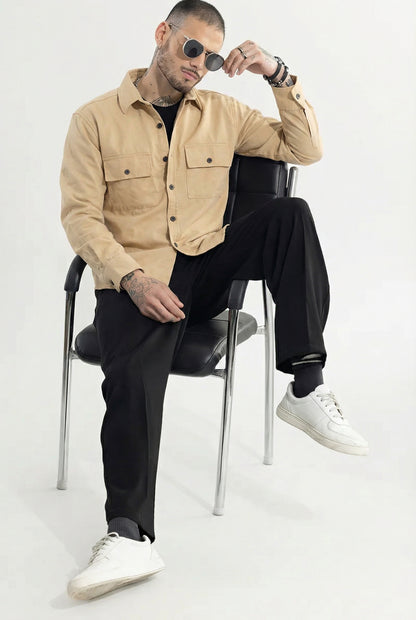 Man in beige button-up overshirt, black pants, white sneakers, and sunglasses sitting on chair