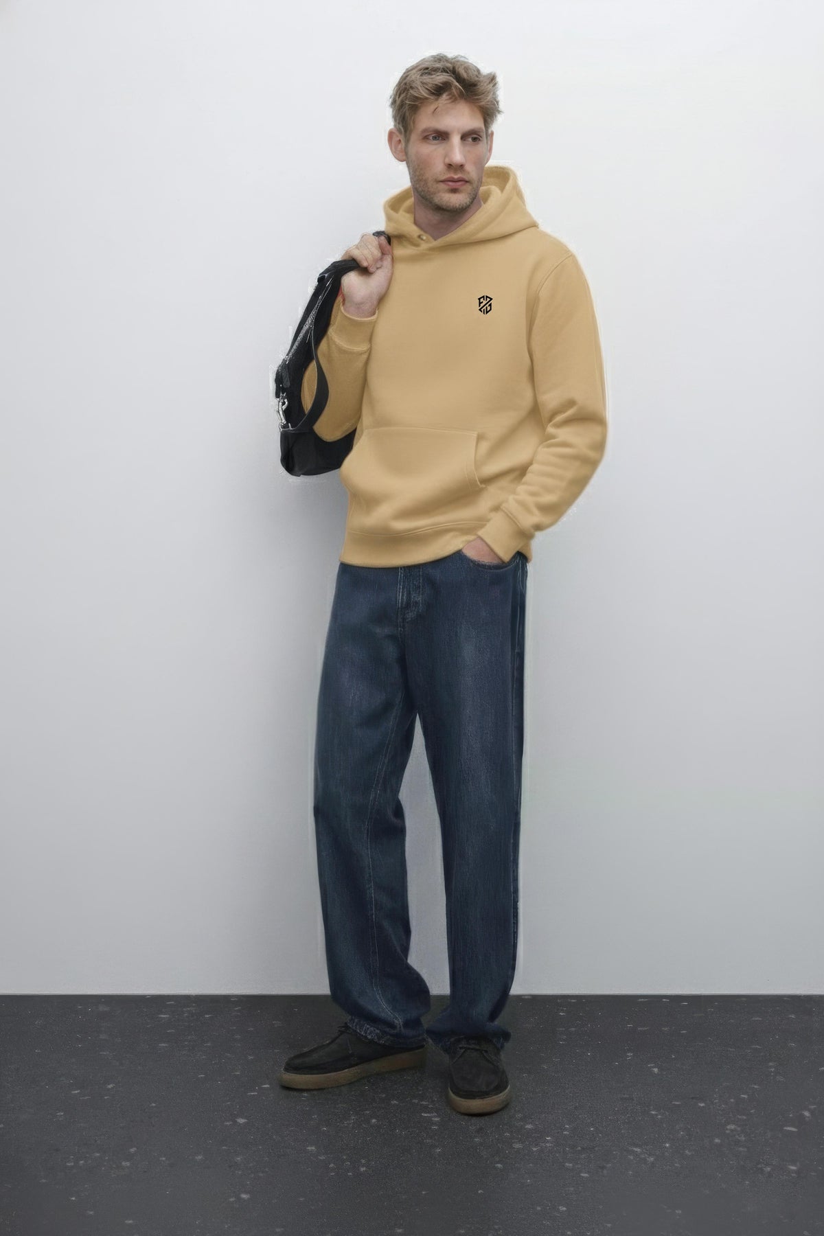 Man wearing tan hoodie with black logo, blue jeans and black sneakers, holding a black bag