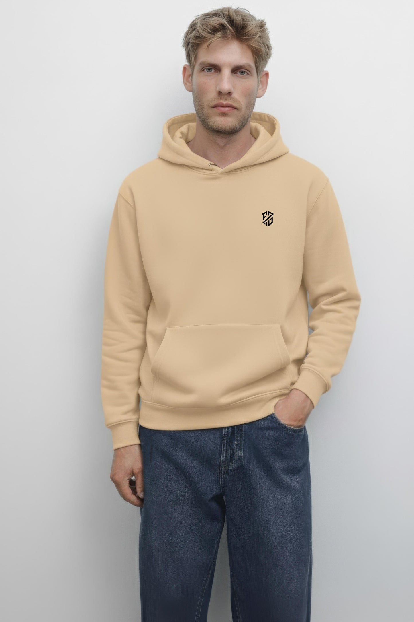 Man wearing beige hoodie with black chest logo and blue jeans, standing against white background