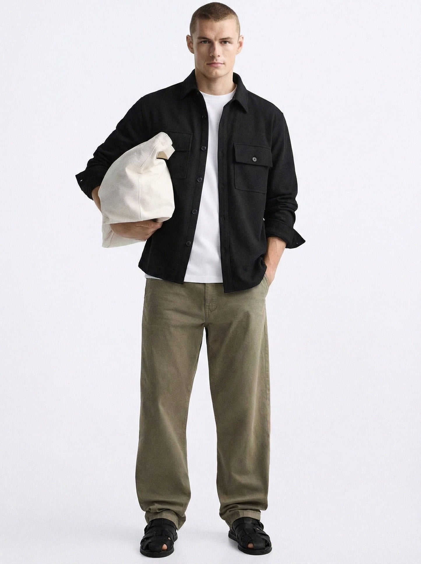 Man in black overshirt, white t-shirt, olive pants, holding cream bag, black sandals