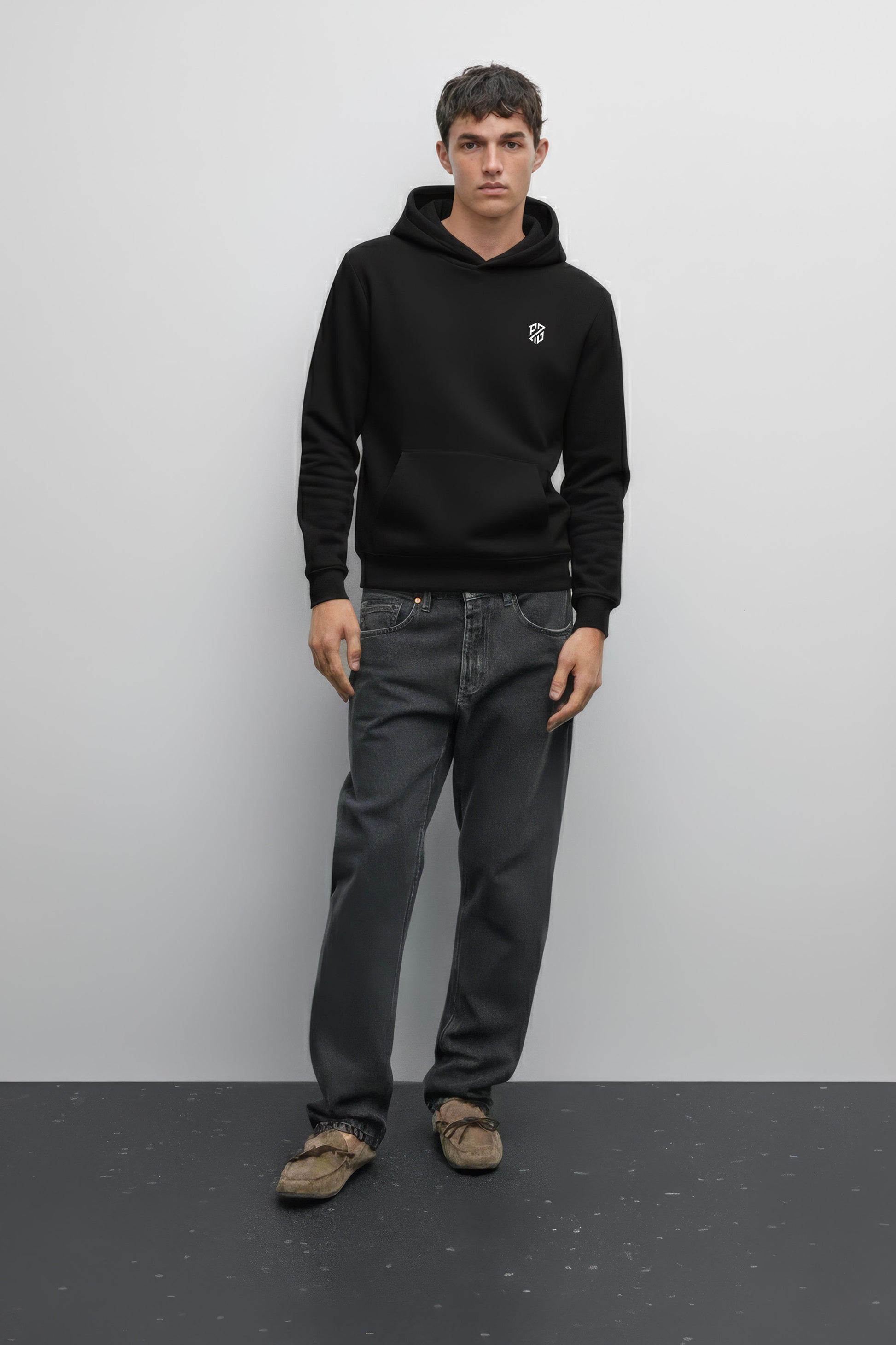 Man in black hoodie with white logo, dark jeans, and brown moccasins standing indoors