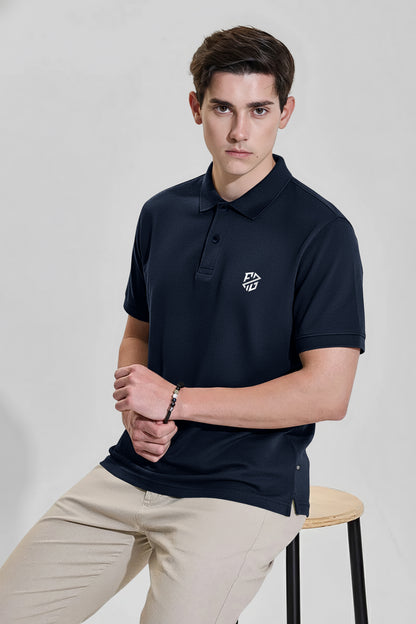 Man wearing a navy blue polo shirt with white logo and beige pants, seated on a stool