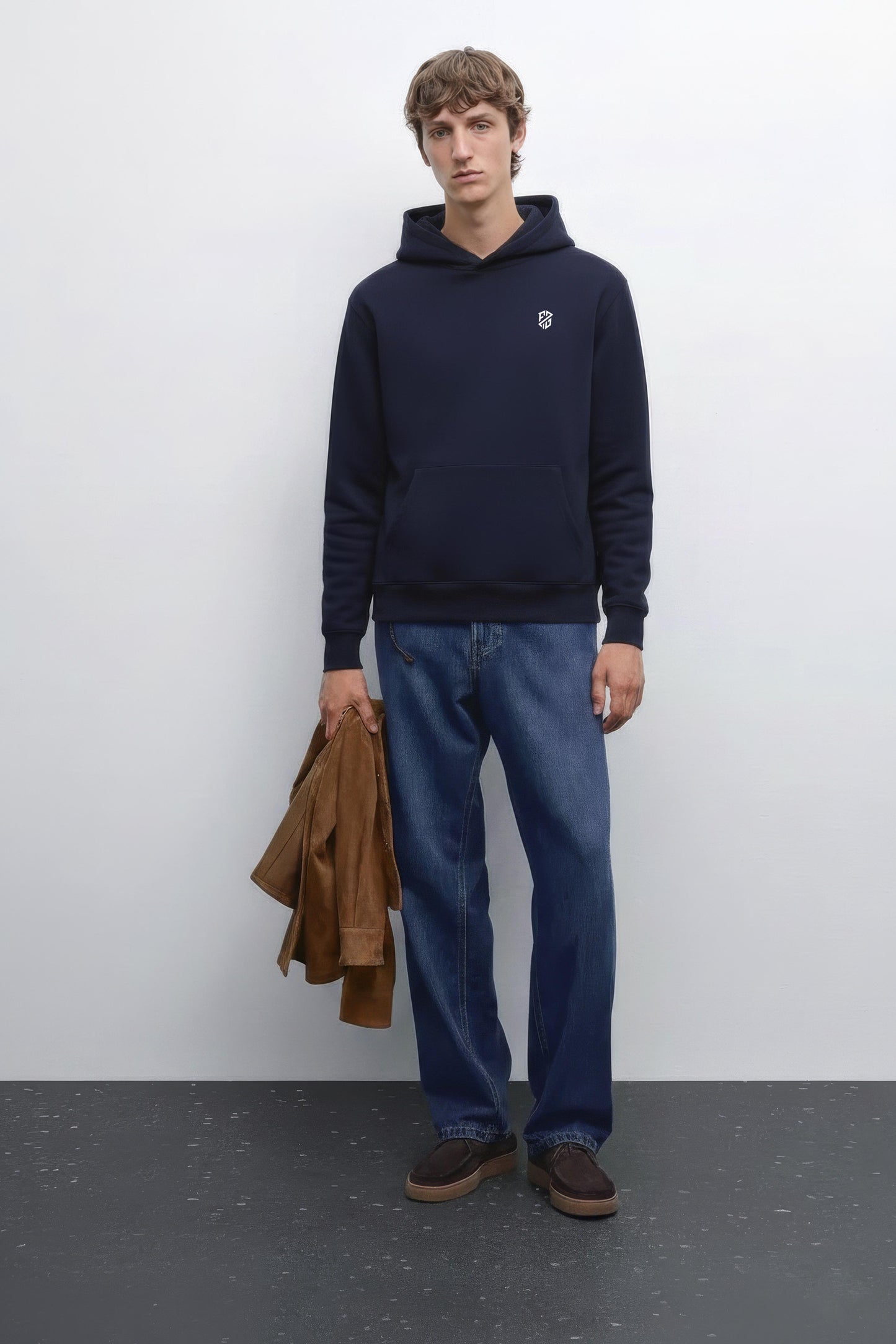 Man in navy hoodie with logo, blue jeans, brown shoes, holding brown jacket