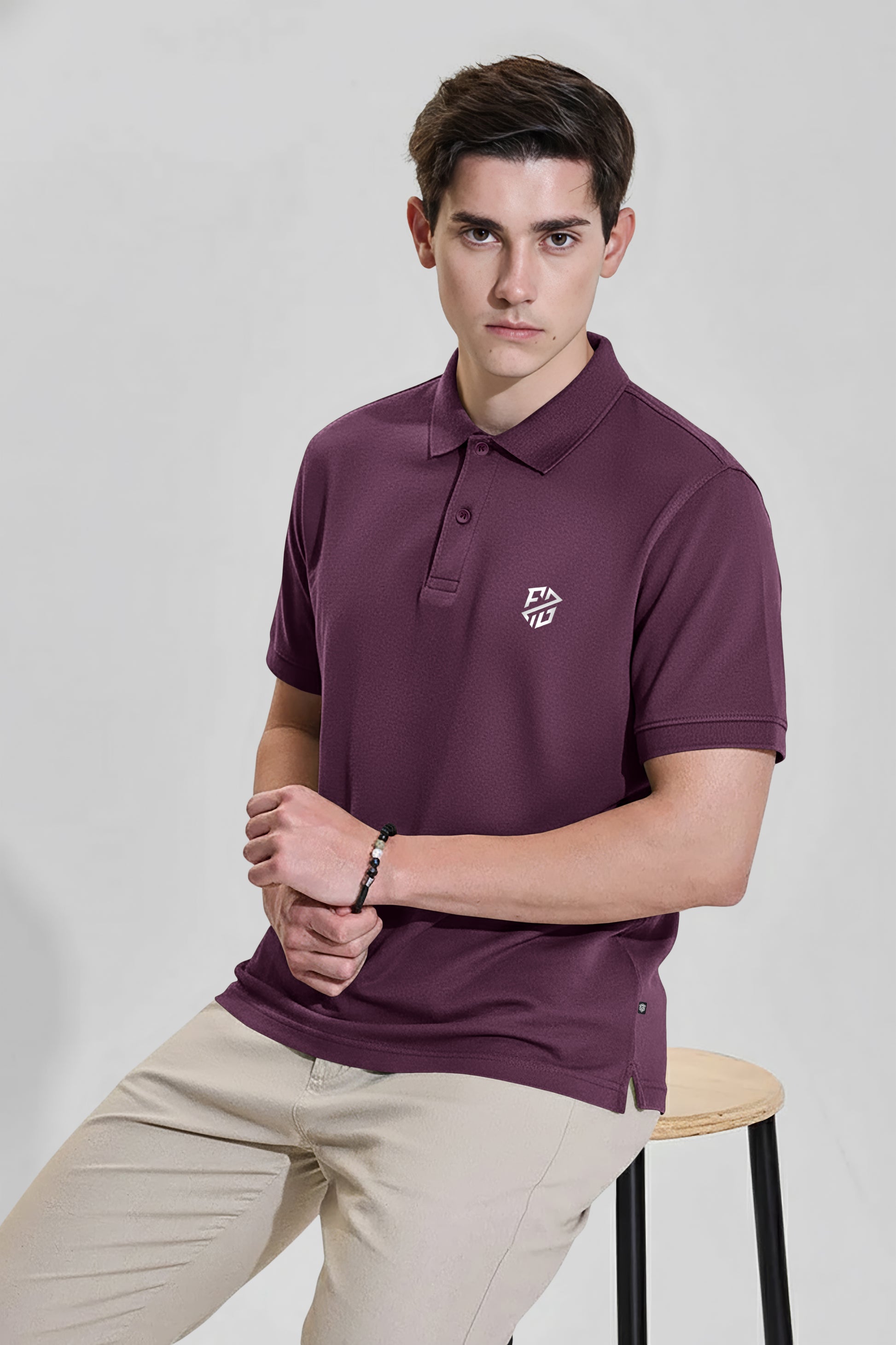 Man wearing maroon short sleeve polo shirt with logo, paired with beige pants, seated on stool