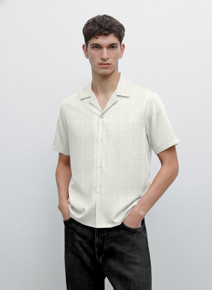 Man wearing short sleeve button-up cream patterned shirt and black jeans, minimalist background