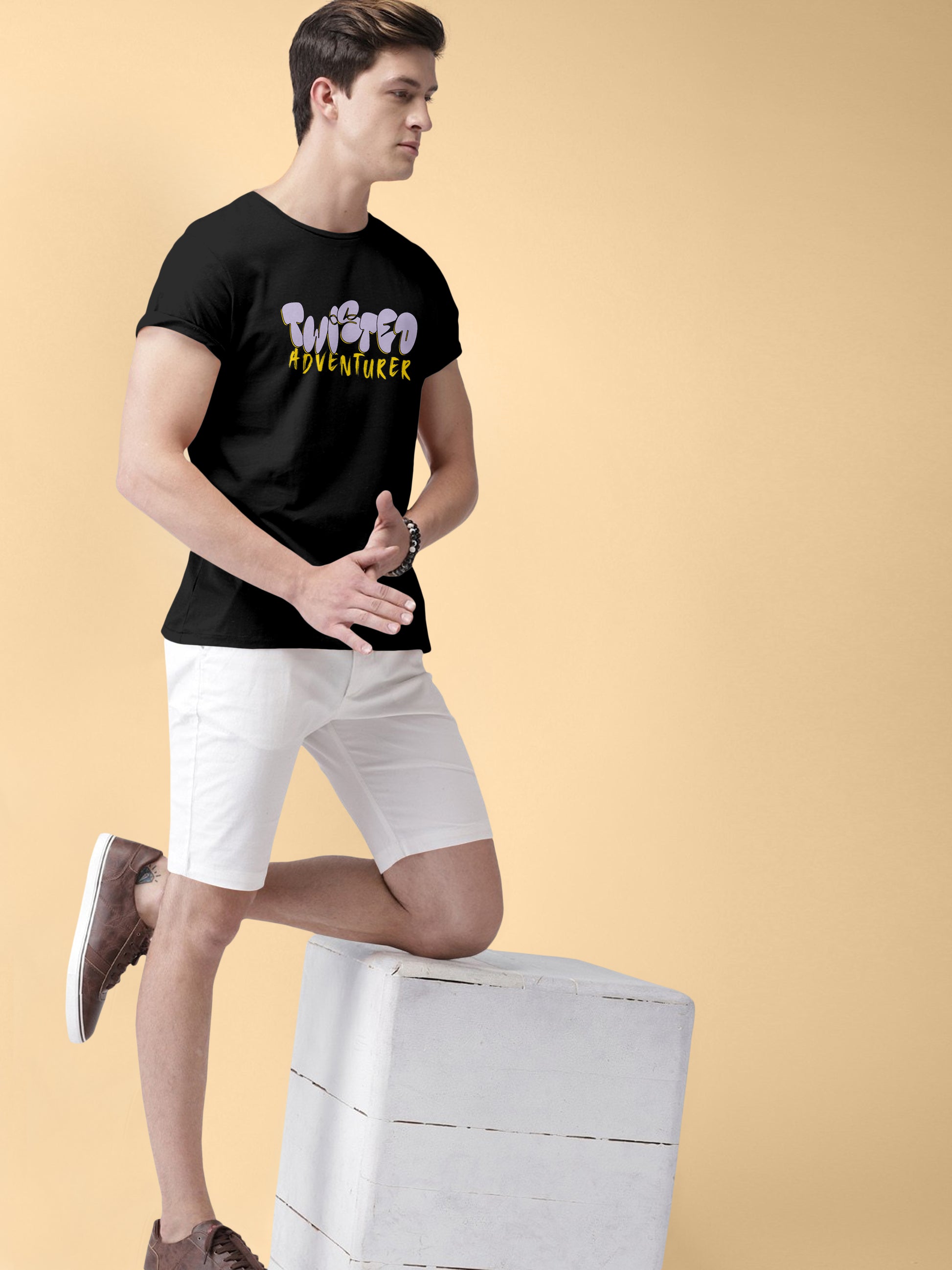 Man wearing black Twisted Adventurer t-shirt, white shorts, brown shoes, posing indoors